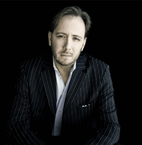 A man with light brown hair and a short beard wears a dark pinstriped suit and white shirt, sitting against a black background.